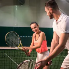 Para z rakietami tenisowymi na korcie, mężczyzna uczy kobietę gry.