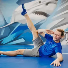 Mężczyzna w niebieskiej koszulce i szarych spodenkach wykonuje ćwiczenie rozciągające nogi na niebieskiej macie.